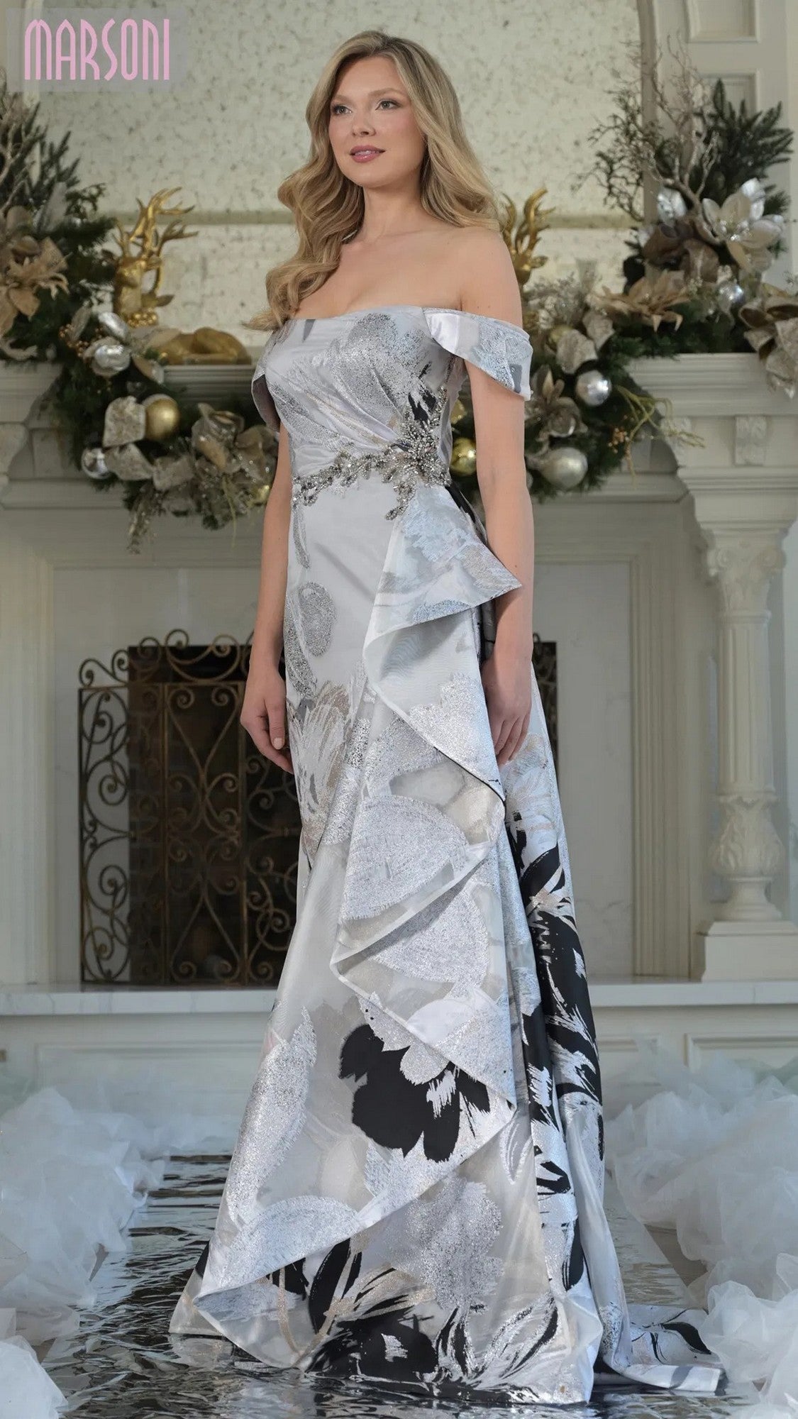 A woman stands indoors near a decorated fireplace wearing the Marsoni MV1368 long formal dress in silver multi.